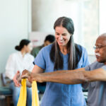 As other therapists work with patients in the background, the mid adult female physical therapist teaches the senior adult man how to use an elastic band to exercise his arms.